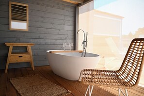 Une salle de bain moderne avec une baignoire sur pieds, un tabouret en bois, un fauteuil en osier et une grande fenêtre donnant sur l’extérieur.