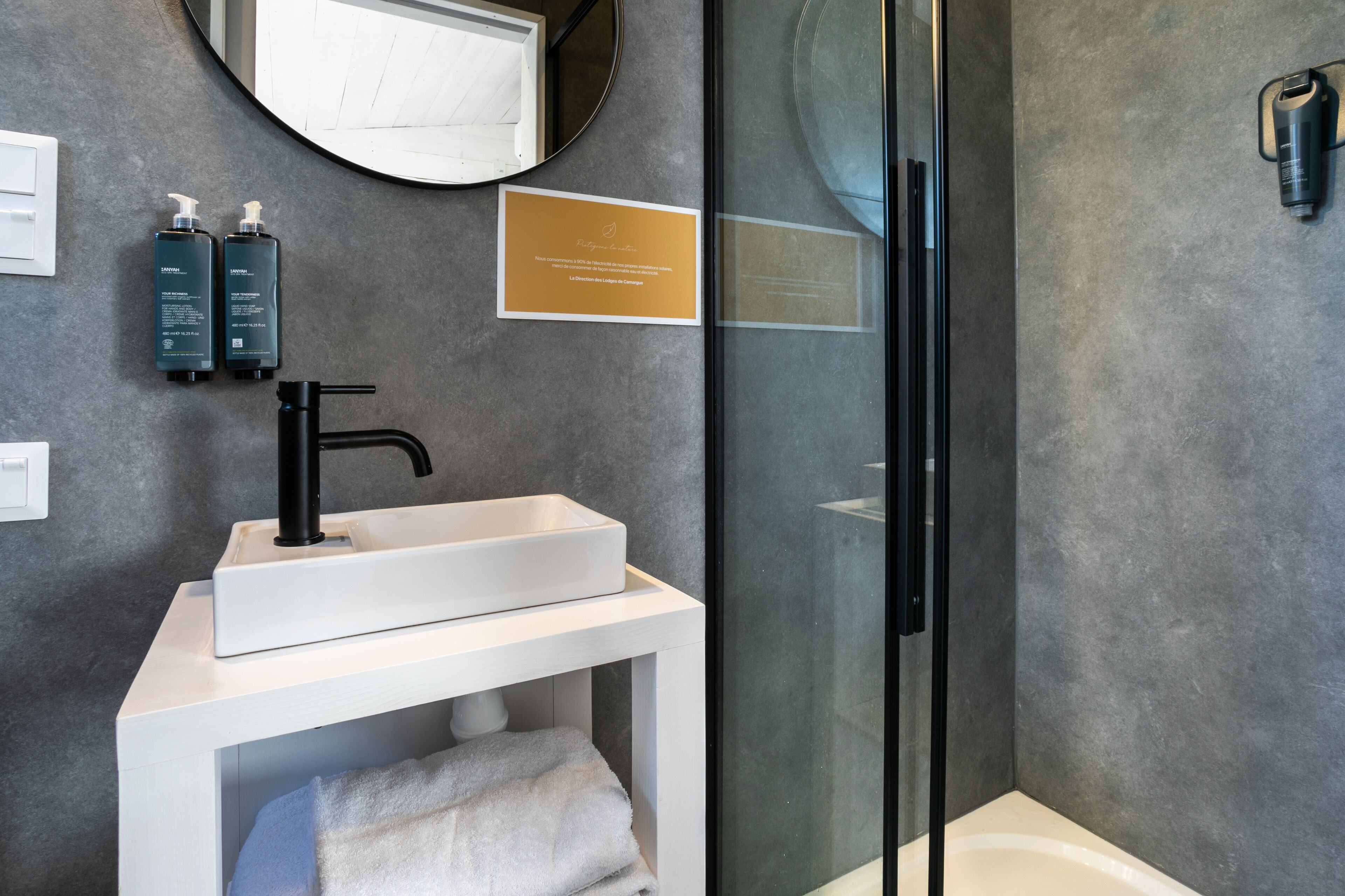 Un baño moderno con un lavamanos blanco, grifo negro y cabina de ducha de vidrio. Se observan dos botellas de champú y una placa en la pared.
