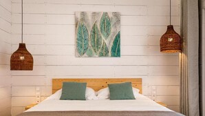 Un lit bien fait, avec une tête de lit en bois, deux oreillers verts et une couverture blanche. Au-dessus du lit, un tableau représentant des feuilles est accroché au mur, et deux lampes suspendues en osier pendent du plafond.