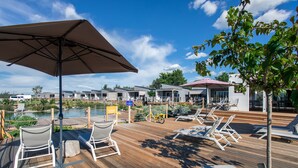 Une terrasse en bois avec des chaises longues et un parasol, donnant sur une piscine et des bâtiments.