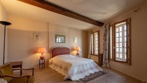 Une chambre à coucher avec un lit, deux lampes de chevet, une chaise, une fenêtre avec des rideaux et une poutre en bois au plafond.
