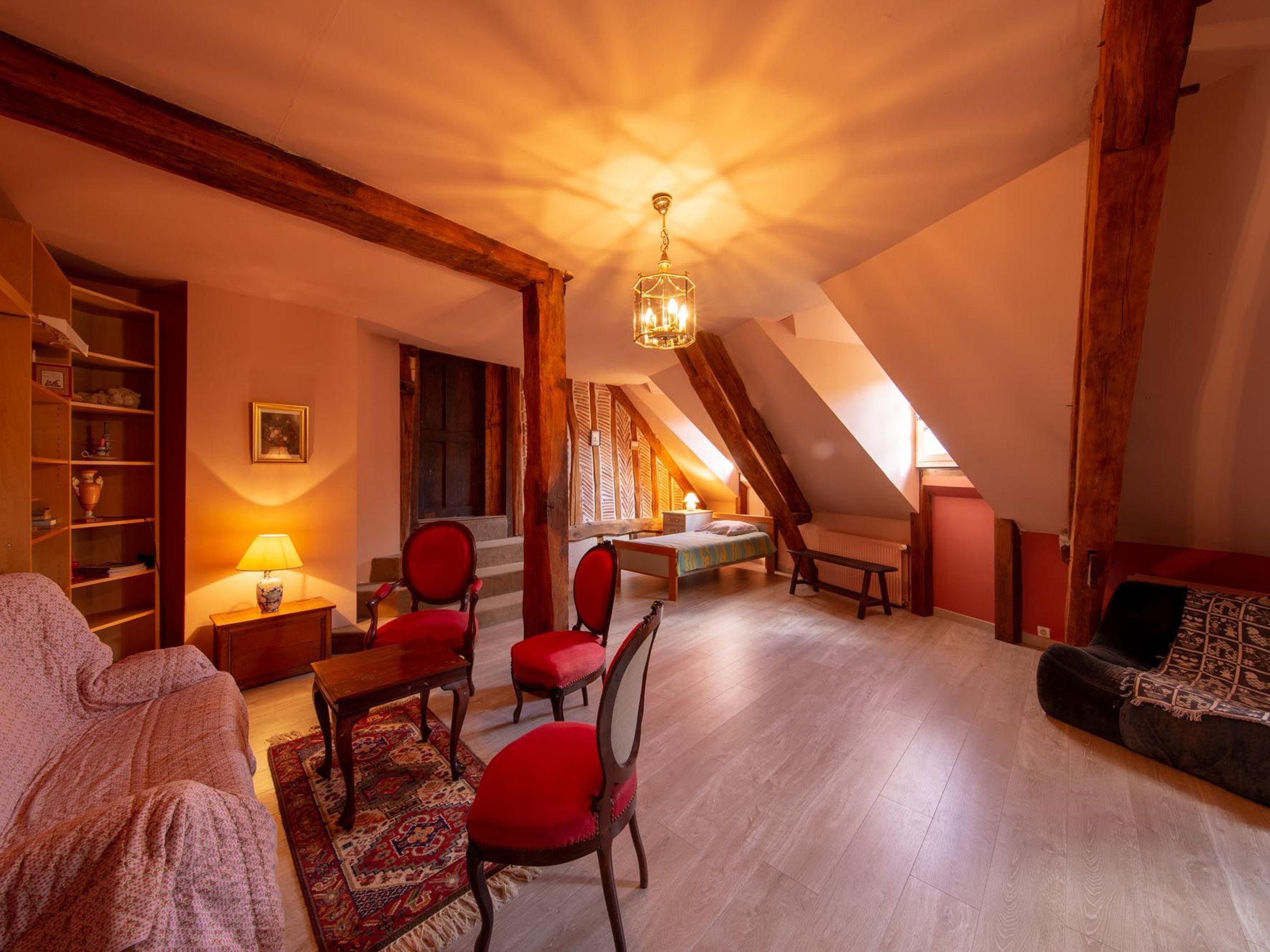 A cozy attic room with a sloped ceiling, wooden beams, a chandelier, a bed, a bench, and a red chair.