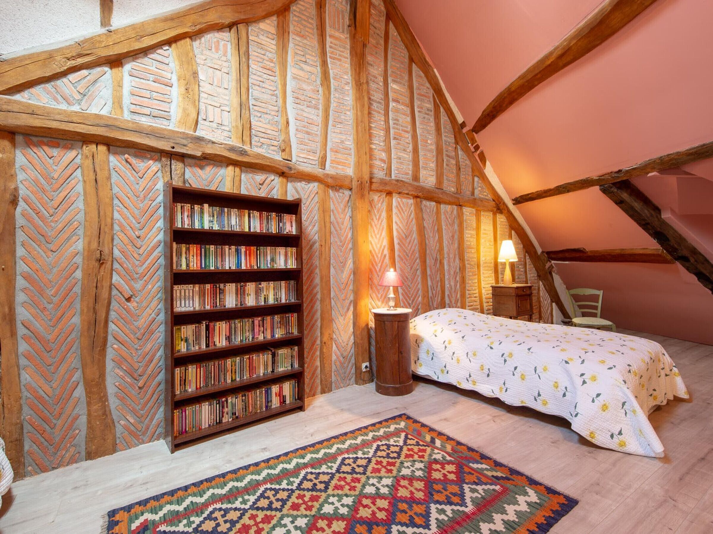 A cozy attic bedroom with exposed wooden beams, a bookshelf, a bed with a floral duvet, and a patterned rug.