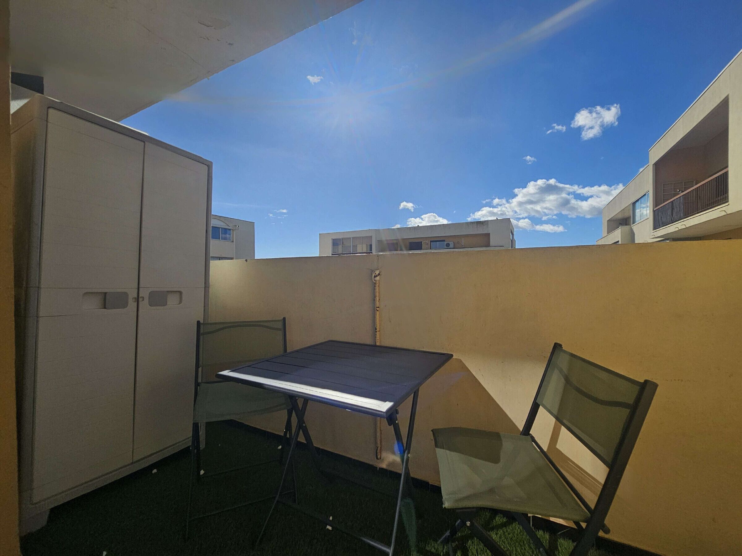 Un pequeño espacio al aire libre con una mesa, sillas, un armario y vistas a edificios bajo un cielo despejado.