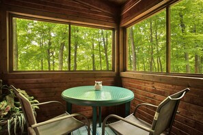 Uma cabana de madeira com uma mesa redonda e duas cadeiras, grandes janelas com vista para uma floresta e um pequeno objeto decorativo sobre a mesa.