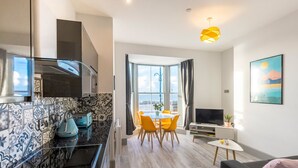 A modern kitchen with a black countertop and patterned backsplash, a dining area with a table and chairs, a sofa, a TV, and a large window with curtains. - Brand new luxury apartment with stunning sea views (Aberystwyth)