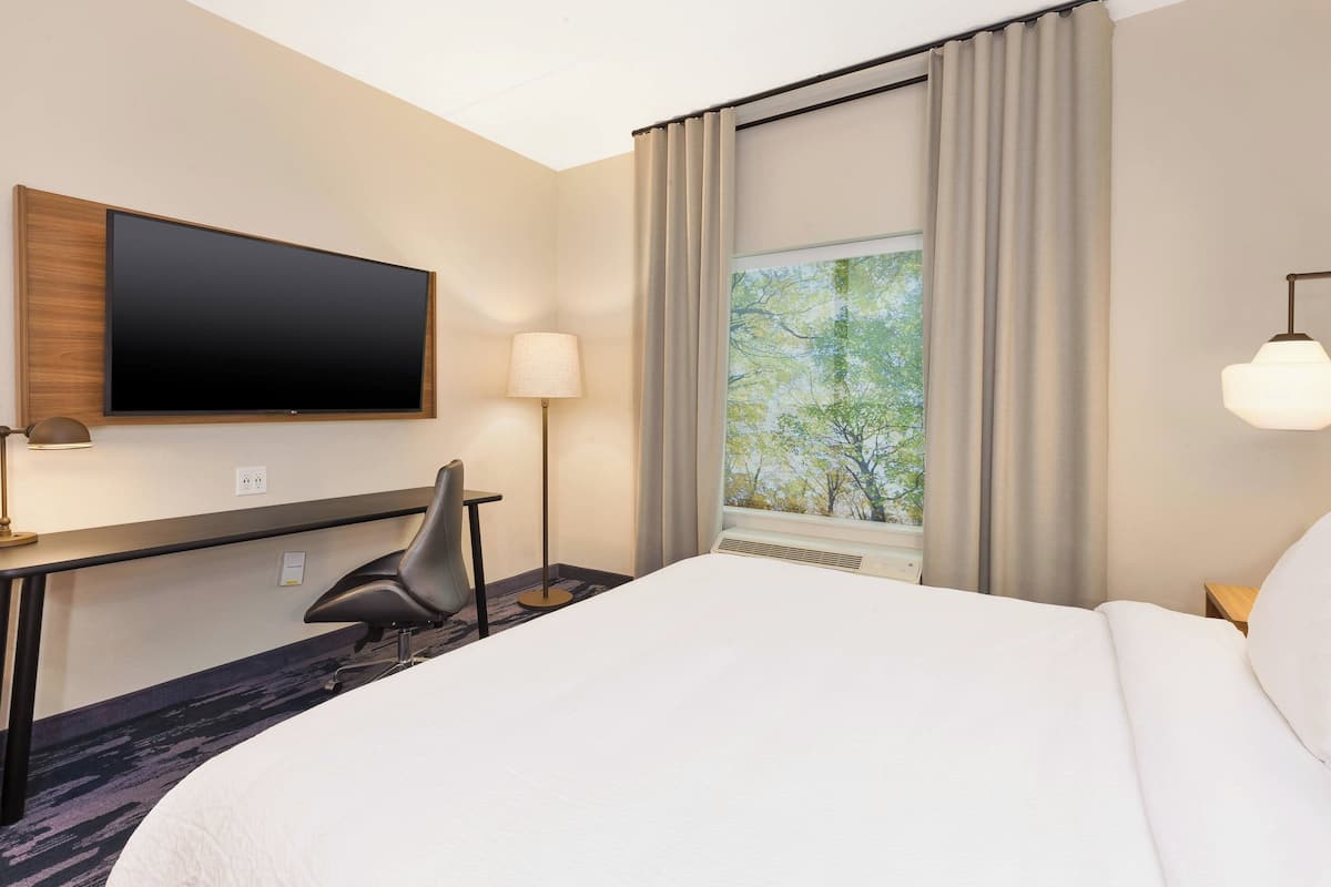 A hotel room with a large bed, a flat-screen TV mounted on the wall, a desk with a chair, and a window with curtains.