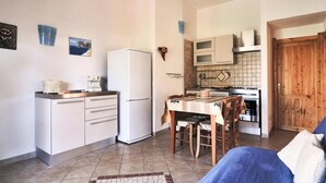 A compact kitchen with a refrigerator, microwave, and stove. There is a dining table with chairs and a blue sofa in the living area. - Apartment on Costa Rei with Sea View (Costa Rei)