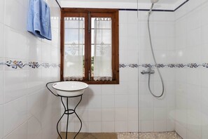 Ein Badezimmer mit weiß gefliesten Wänden, einem Holzkastenfenster mit dekorativen Vorhängen, einem weißen Waschbecken auf schwarzem Fuß und einer Dusche mit Handbrause.