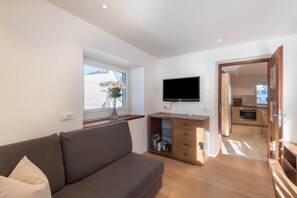 A modern living room with a sofa, a flat-screen TV mounted on the wall, a wooden desk with drawers, and a window with a view of a snowy landscape.