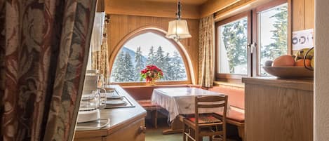 A cozy cabin interior with a dining area, wooden walls, a large window with a mountain view, and a table with chairs.