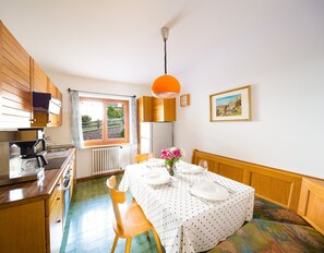 Een keuken met een kookplaat, spoelbak en raam. Er staat een eettafel met stoelen en een bloemenvaas.
