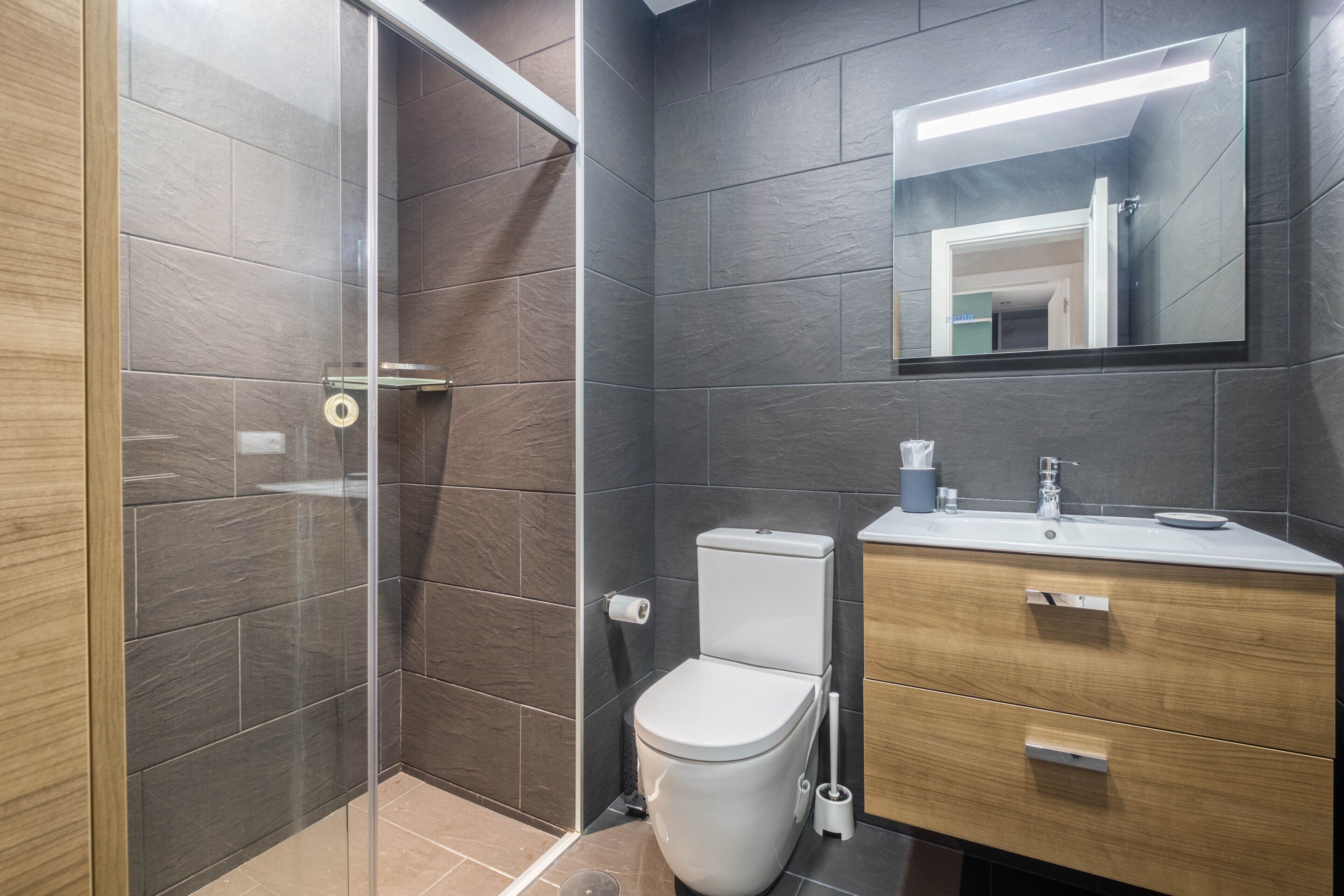 Une salle de bain moderne avec une douche à parois de verre, des toilettes blanches, un meuble-lavabo en bois avec un miroir et un évier.