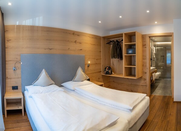 Una habitación de hotel moderna con cabecera de madera, dos camas con ropa blanca, un armario empotrado y zona de ducha.