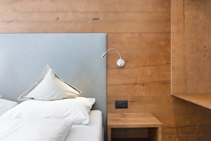A bed with white linens and pillows, a wooden nightstand with a power outlet, and a wall-mounted light fixture.
