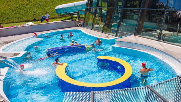 Une piscine avec plusieurs couloirs de nage, un toboggan aquatique et un bâtiment en verre en arrière-plan.
