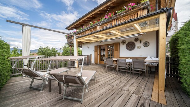 Una terraza de madera con mobiliario exterior, un balcón con flores y vistas a la montaña.
