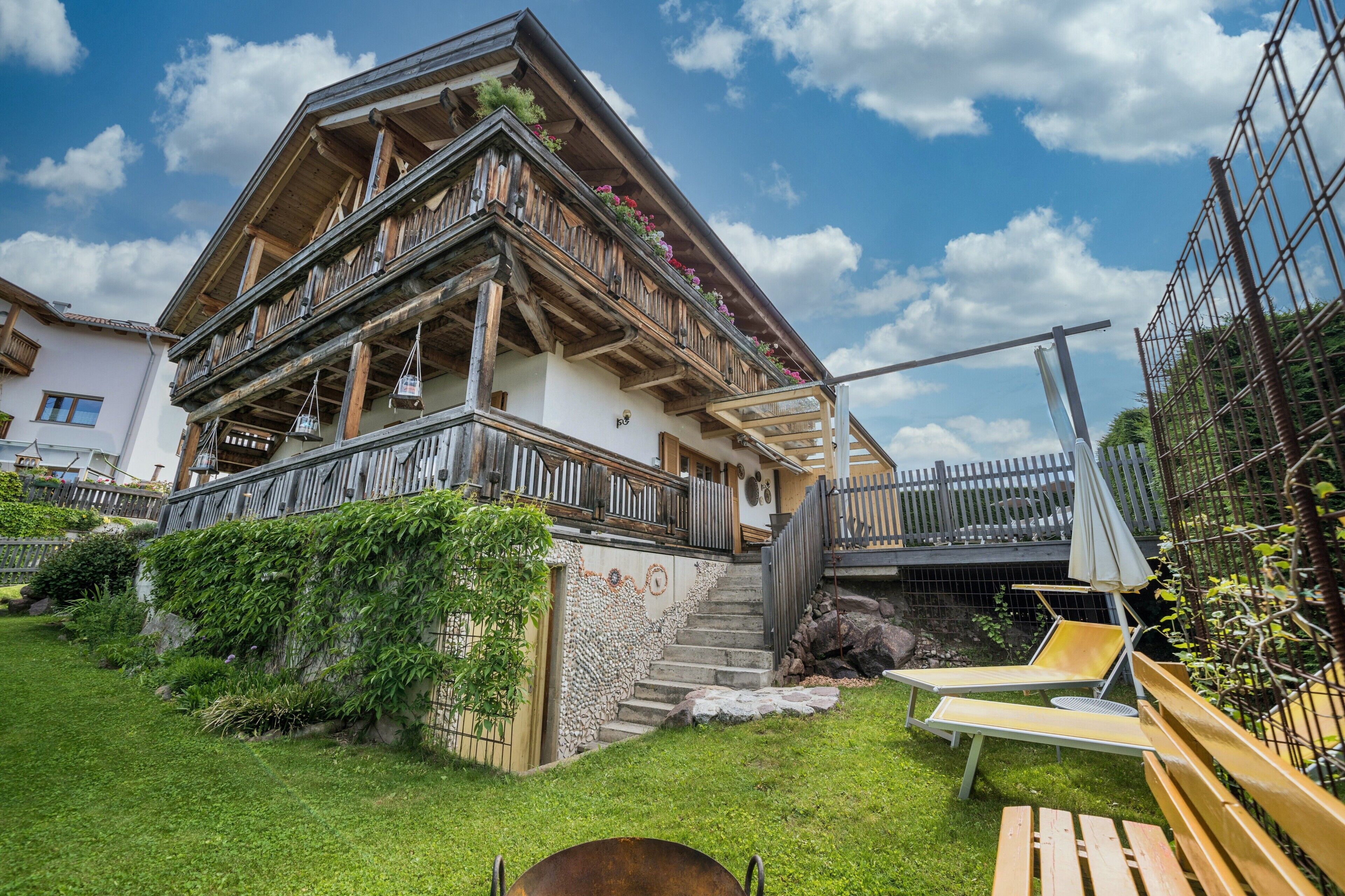 A traditional chalet with a wooden balcony, a stone staircase, and a sunny outdoor seating area.