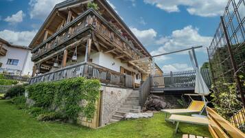 A traditional chalet with a wooden balcony, a stone staircase, and a sunny outdoor seating area.