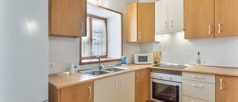 A kitchen with wooden cabinets, a white refrigerator, a microwave, a sink, and a window with curtains.