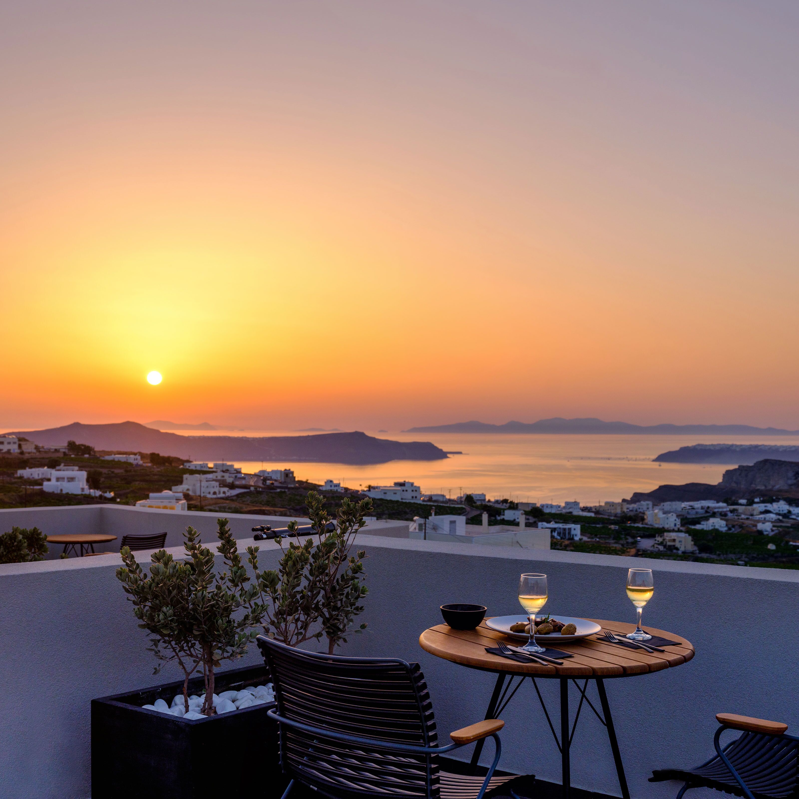 Une terrasse avec une table dressée pour deux personnes, offrant une vue sur un paysage côtier au coucher du soleil.