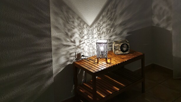 Ein Zimmer mit einem Holztisch, einer dekorativen Leuchte, die Schatten wirft, und kleinen gerahmten Bildern an der Wand.