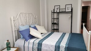 Ein ordentlich gemachtes Bett mit einem gemusterten Bettbezug und Kissen, ein weißes Metall-Kopfteil, ein kleiner Nachttisch mit einer grünen Flasche und einem Dekorationsgegenstand sowie ein schwarzes Metallregal mit gerahmten Bildern an der Wand.