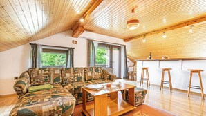 Una sala de estar acogedora con techo de madera, un sofá estampado, una mesa de centro y una zona de cocina con taburetes.
