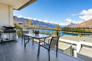 A balcony with a table and chairs, a grill, and a view of mountains and a lake.