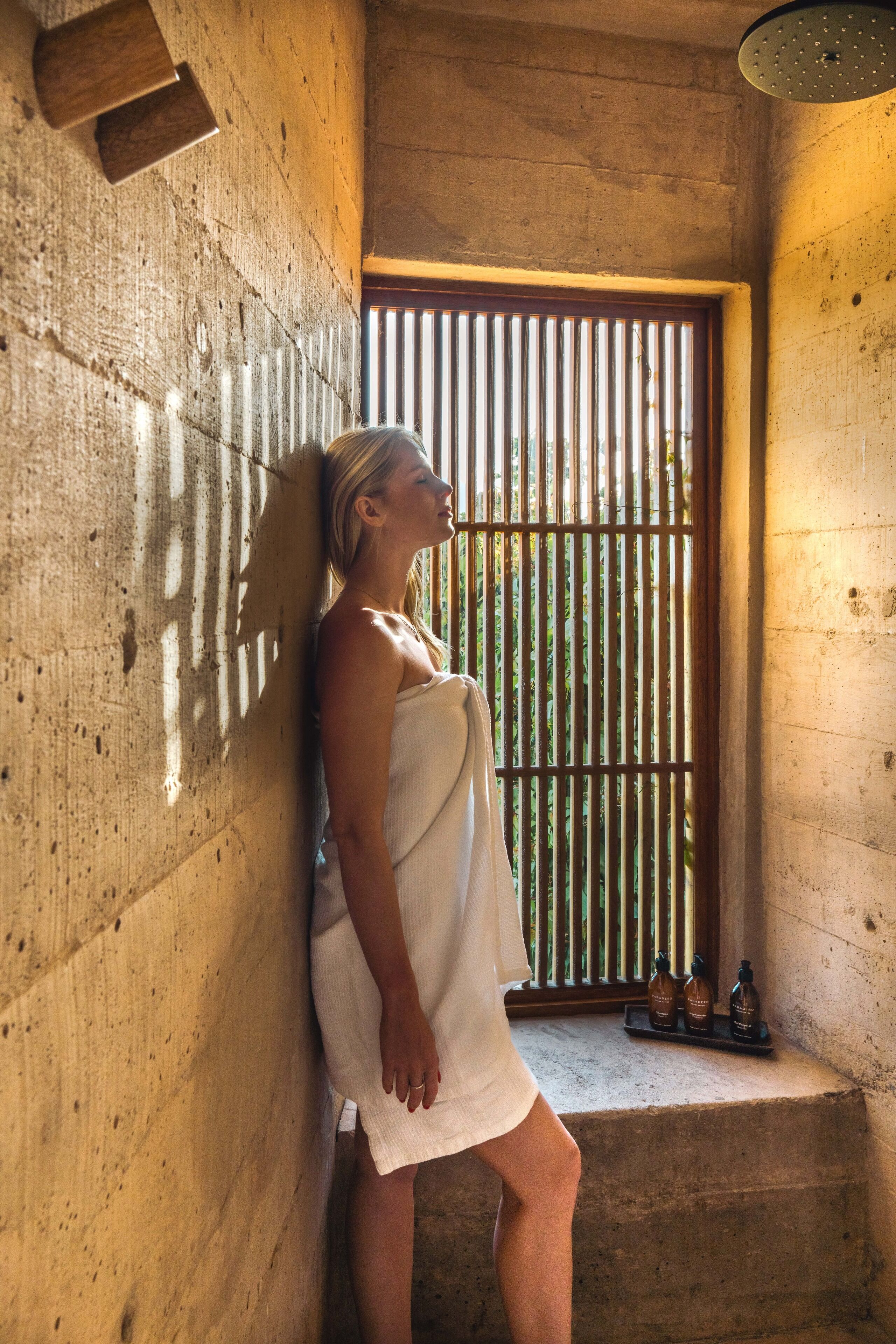 A person in a towel leaning against a wooden wall in a room with a window and a shelf with bottles.
