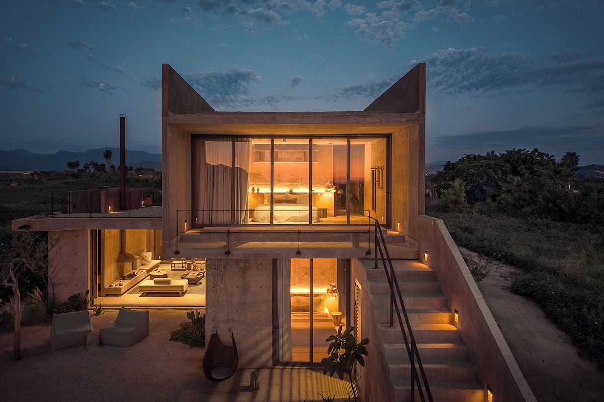 Una casa moderna con un amplio ventanal que da a un dormitorio, una escalera y un patio con mobiliario.