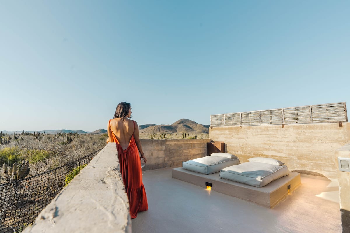 Una mujer con un vestido rojo está de pie en una terraza en la azotea con vistas a un paisaje desértico y montañas a lo lejos.