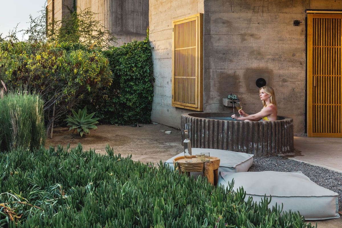 Un edificio moderno con fachada de concreto, amplias ventanas y un pequeño espacio al aire libre con cojines y una mesa.