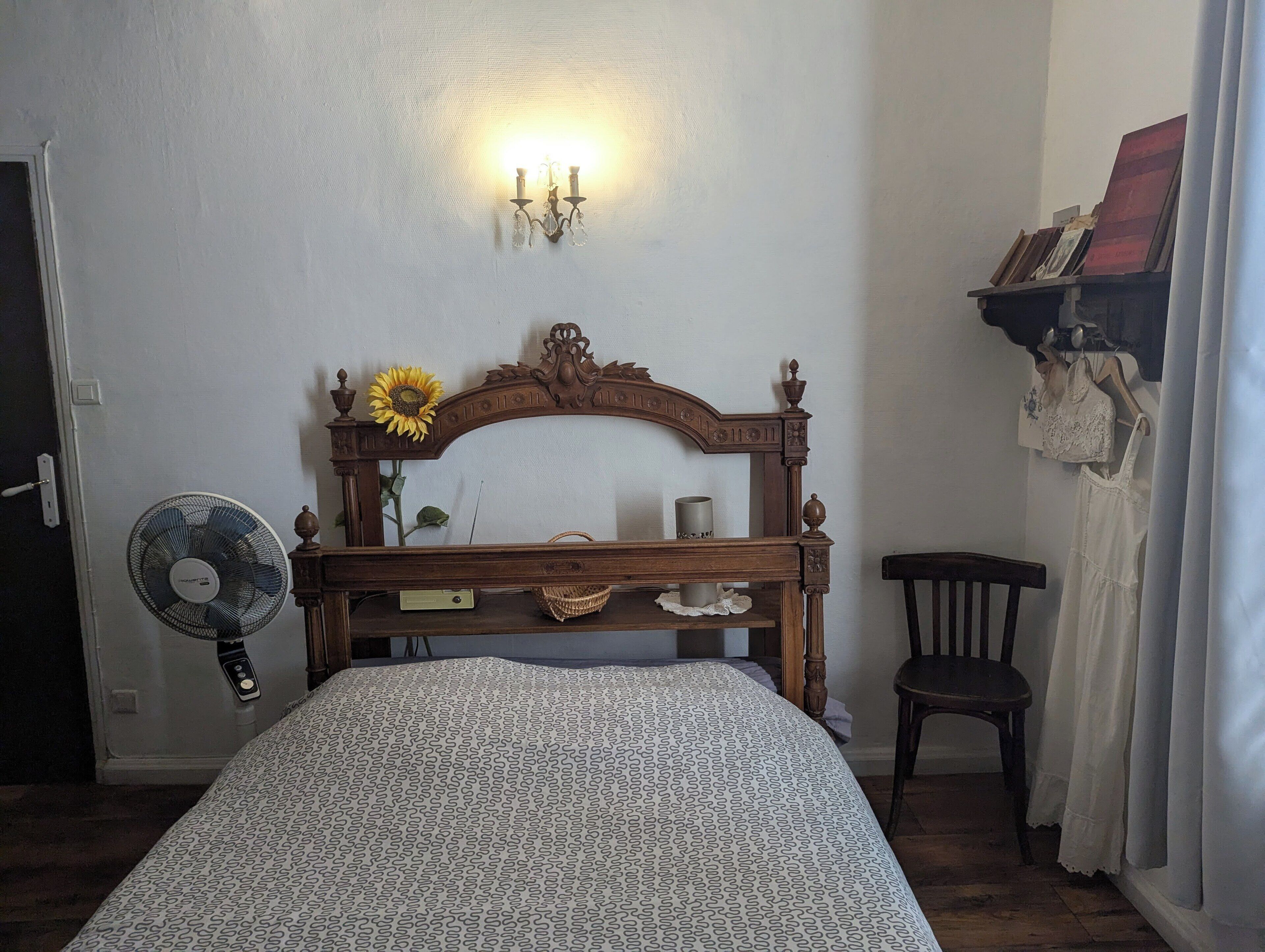 Een slaapkamer met een houten bed, een ventilator, een wandlamp en een plank met kleding.