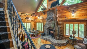 A log cabin interior with a stone fireplace, wooden floors, a staircase, and a dining area.