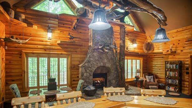 A log cabin interior with a stone fireplace, wooden furniture, and hanging light fixtures.