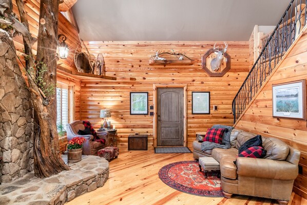 A log cabin interior with a stone fireplace, wooden walls, a leather sofa, a round rug, and a staircase. - Iron Mountain Lodge 3 Bedroom Cabin by RedAwning (Butler)