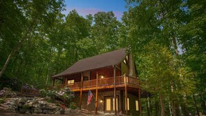 A two-story log cabin with a balcony, surrounded by dense forest.