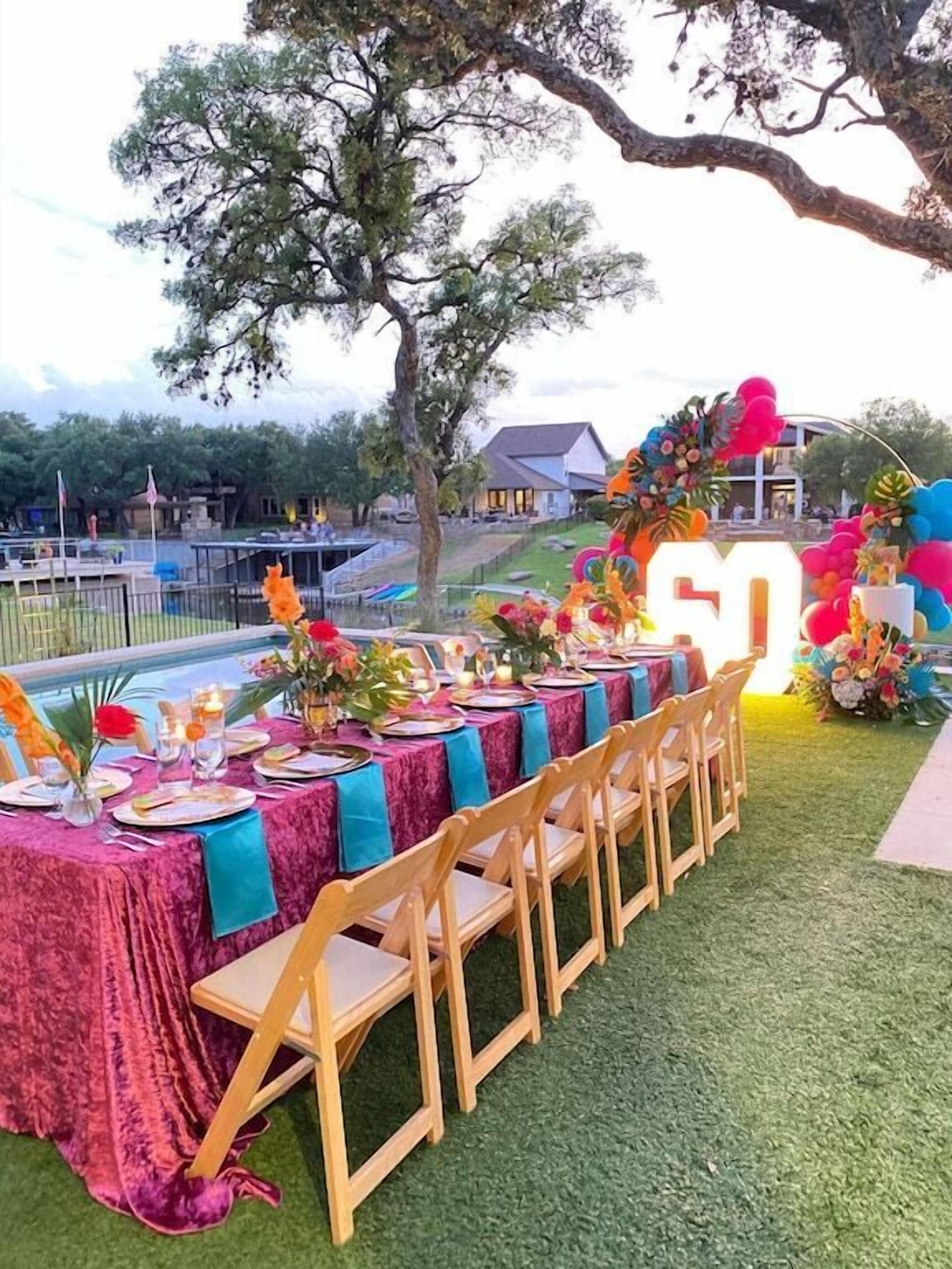 An outdoor dining area with a long table set for a meal, surrounded by chairs and decorated with flowers and balloons.