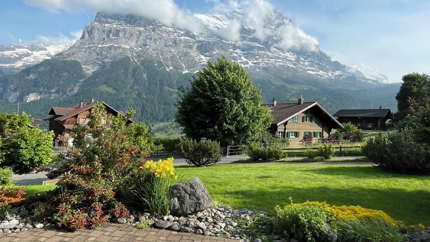 A mountain landscape with snow-capped peaks and a valley with houses and greenery.