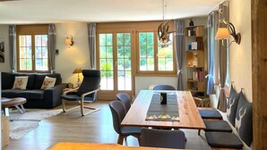 A spacious living room with a wooden dining table, comfortable seating, and large windows offering a view of greenery.