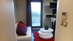 Een moderne kamer met een witte bank, rode kussens, een kleine plant op een witte ronde tafel, een rood vloerkleed, een televisie aan de muur en een raam met uitzicht op bomen.