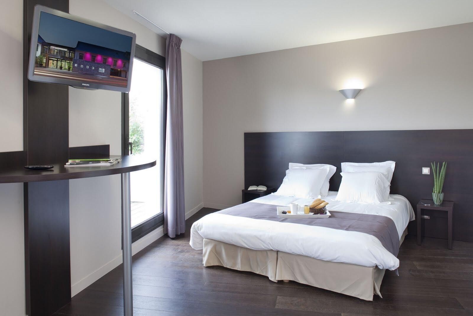 Moderni hotellihuone, jossa on suuri sänky, yöpöytä, seinälle asennettu televisio ja ikkuna, jossa on verhot.