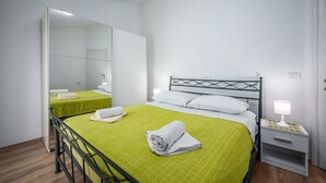 Een eenpersoonsbed met een groene sprei, twee witte kussens, een nachtkastje met een lamp en een grote spiegel.