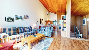 A cozy living room with a wooden ceiling, a sofa set, a coffee table, and a dining area with a kitchen in the background.