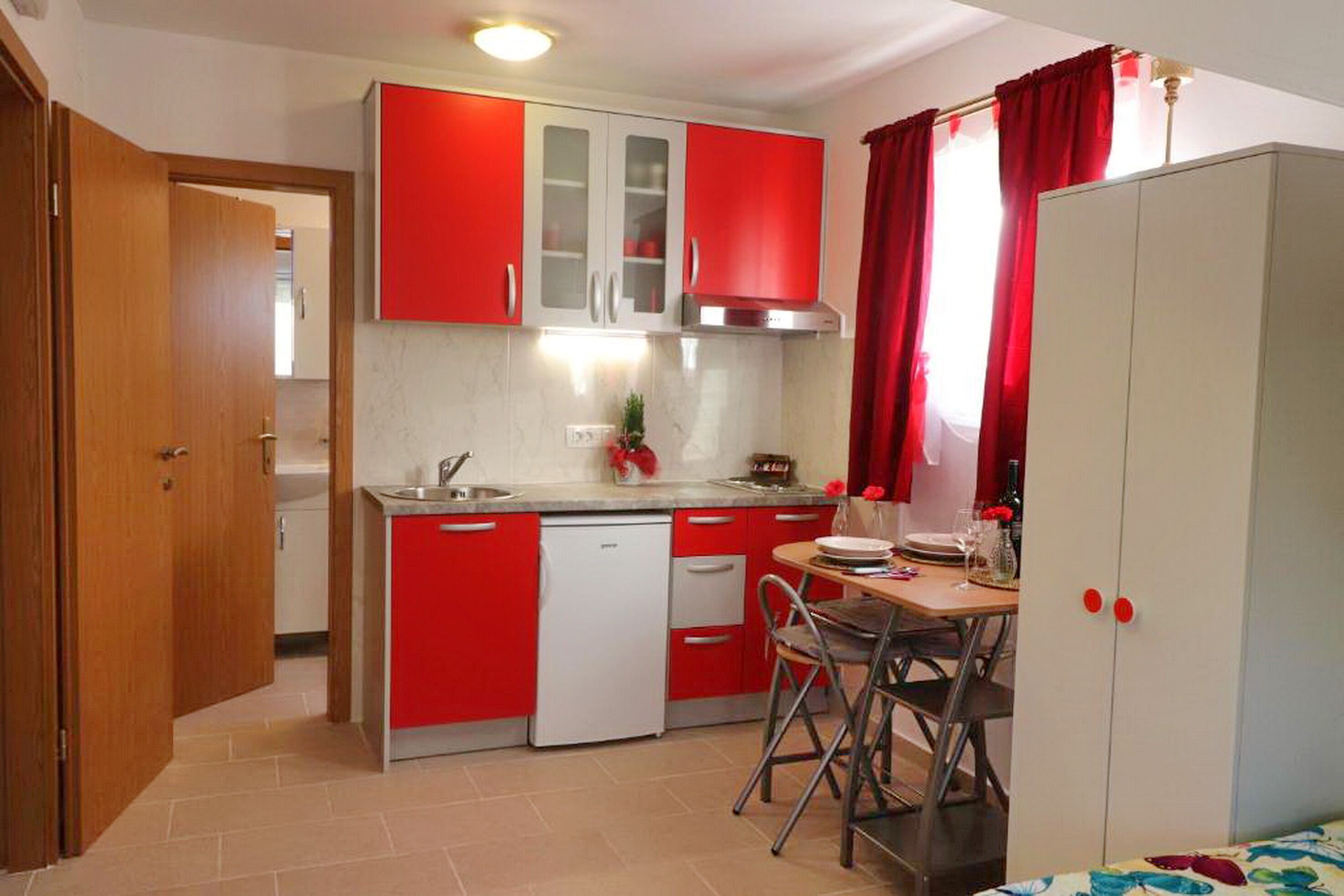 Cocina compacta con armarios rojos, lavavajillas y comedor con mesa y sillas.