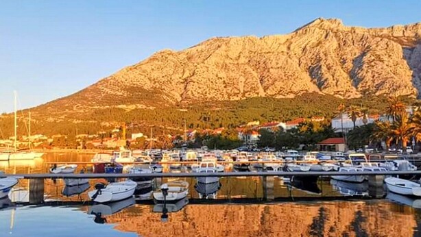 A marina with boats docked in calm waters, reflecting the surrounding landscape and a mountain in the background. - Apartments Clementina Sunrise (Orebic)