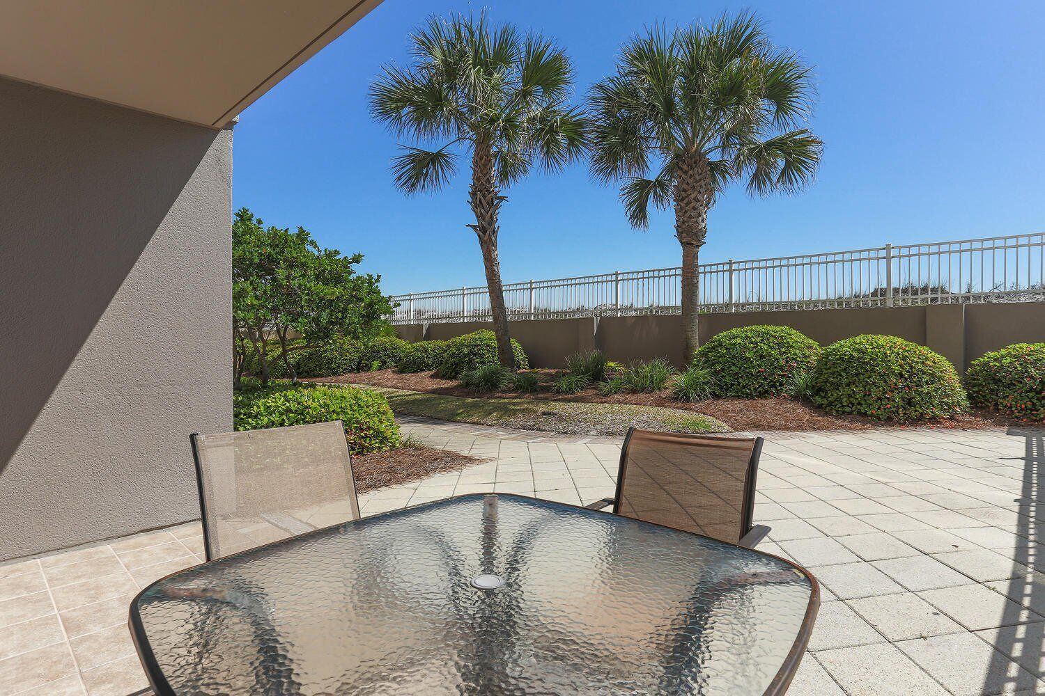 An outdoor patio with a glass table and chairs, surrounded by palm trees and well-maintained bushes.