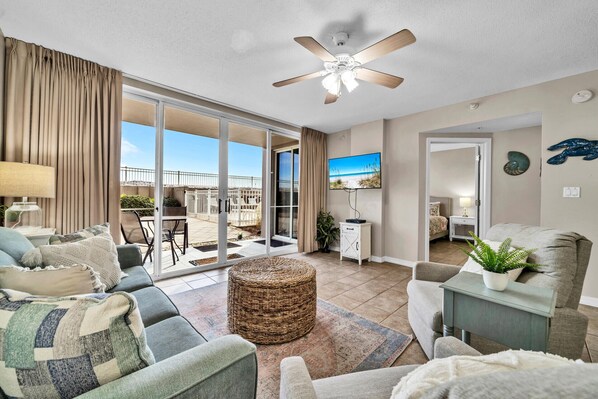A spacious living room with a large sliding glass door leading to a balcony, a ceiling fan, a flat-screen TV mounted on the wall, and a cozy seating area with a woven ottoman.