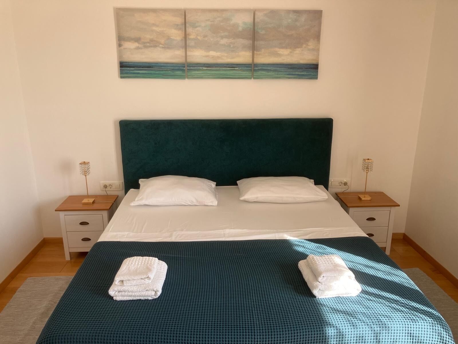 A neatly made bed with a teal headboard, white pillows, and a teal bedspread. There are two bedside tables with lamps, a painting above the bed, and a small wooden nightstand with a lamp.
